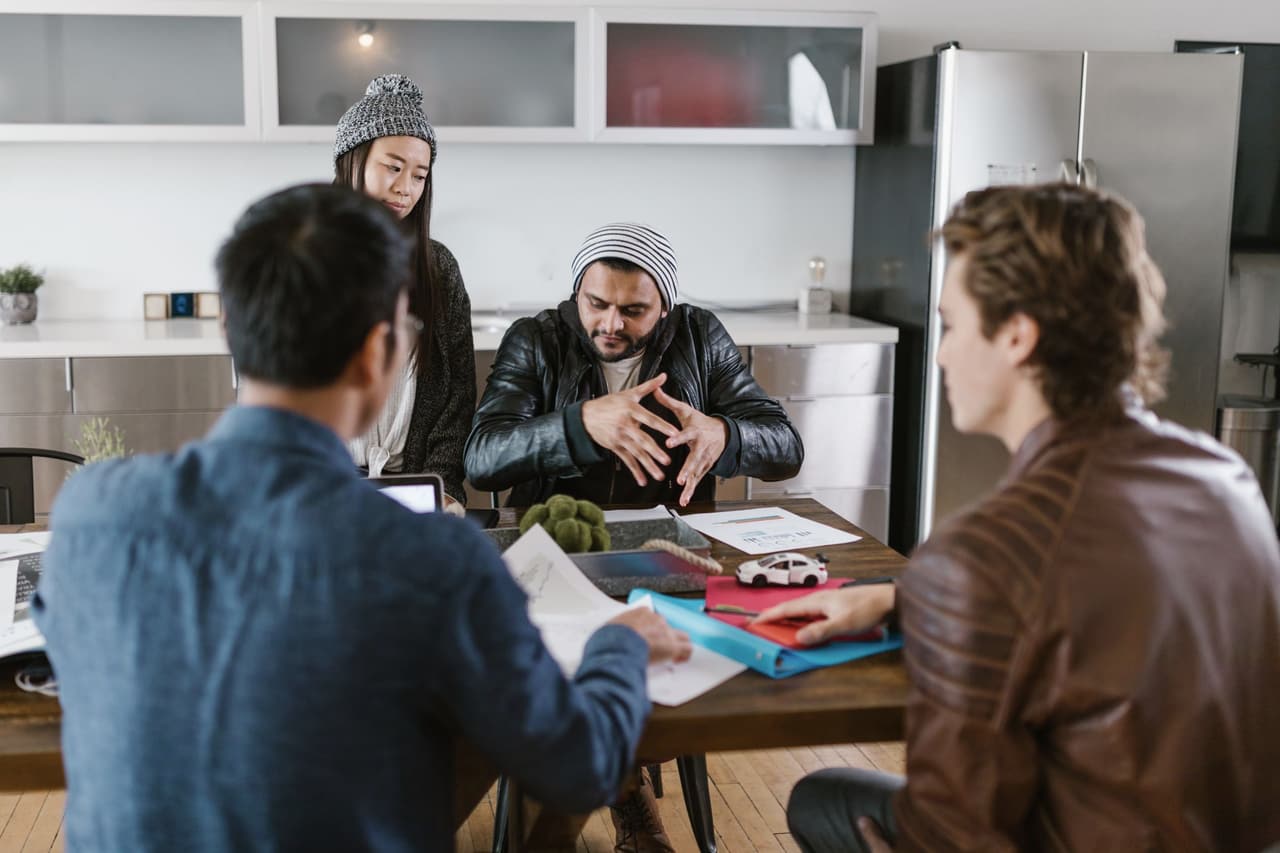 A team in conversation around a worktable in a studio with shelving and large windows