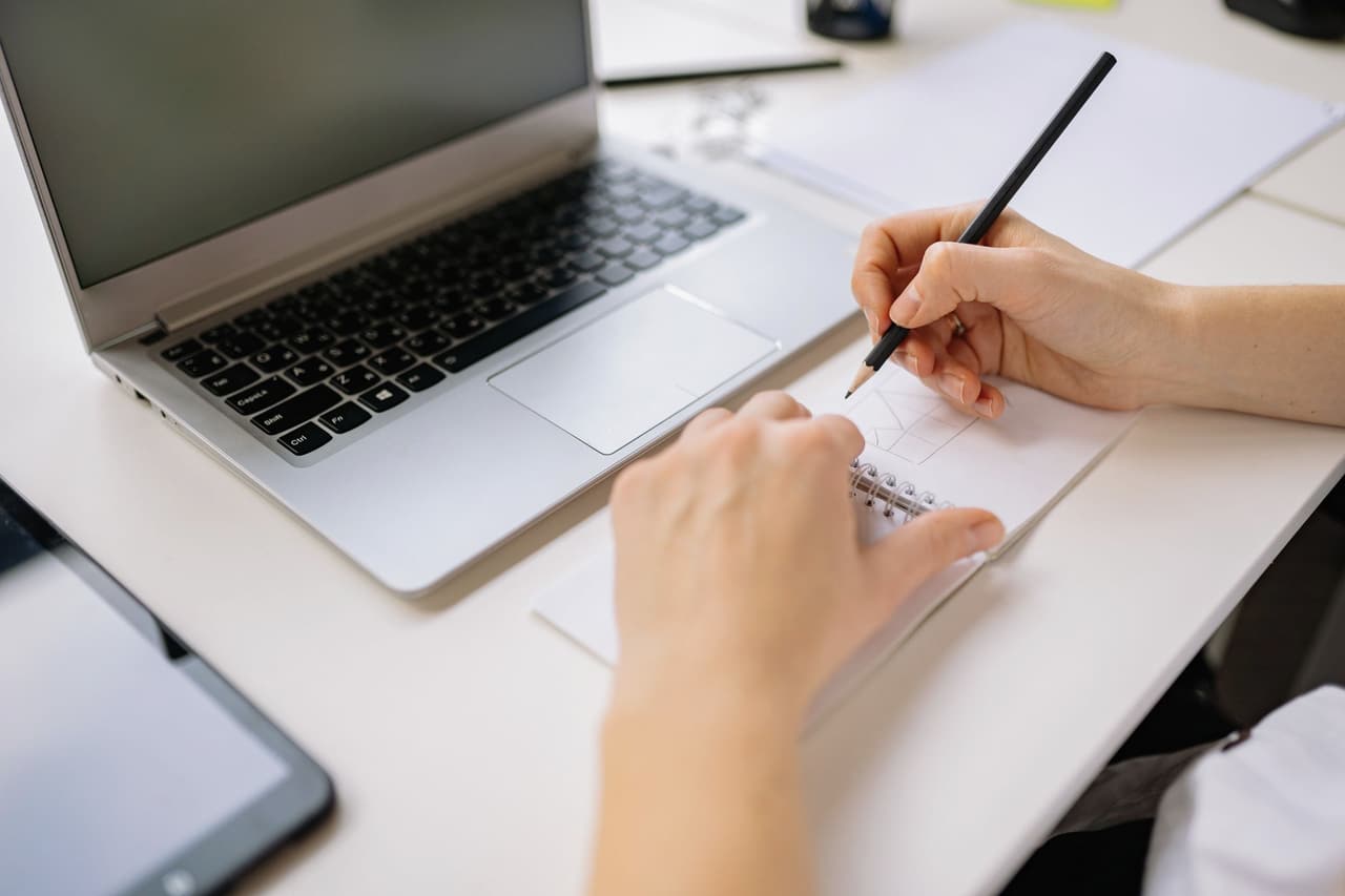A hand sketching in a notebook next to a laptop